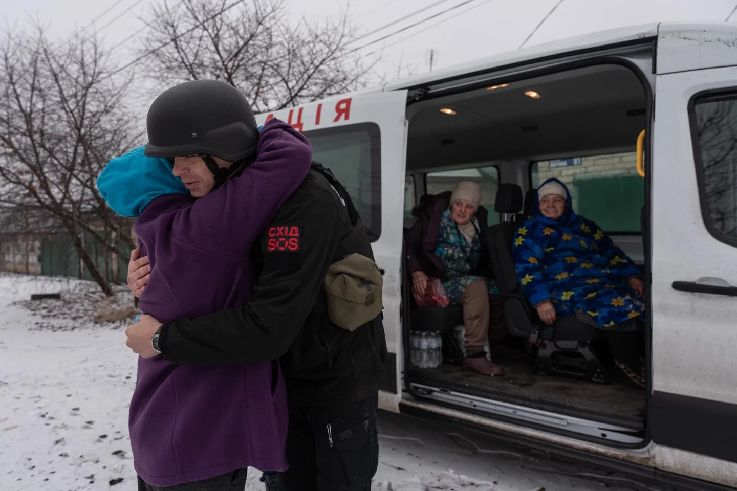 Волонтер у шоломі обіймає жінку біля мікроавтобуса з евакуйованими літніми людьми взимку