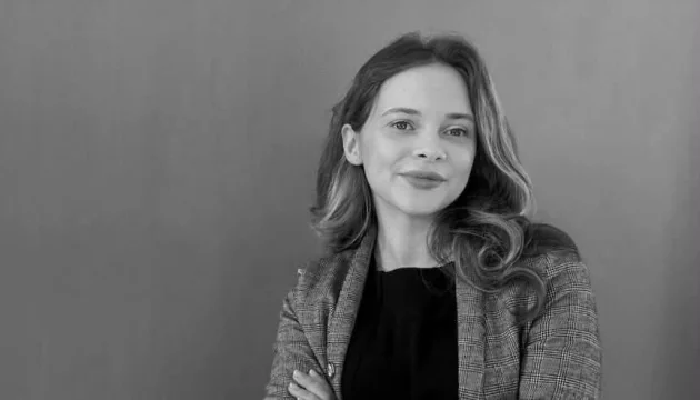Чорно-білий портрет усміхненої жінки з хвилястим волоссям у картатому піджаку зі схрещеними руками